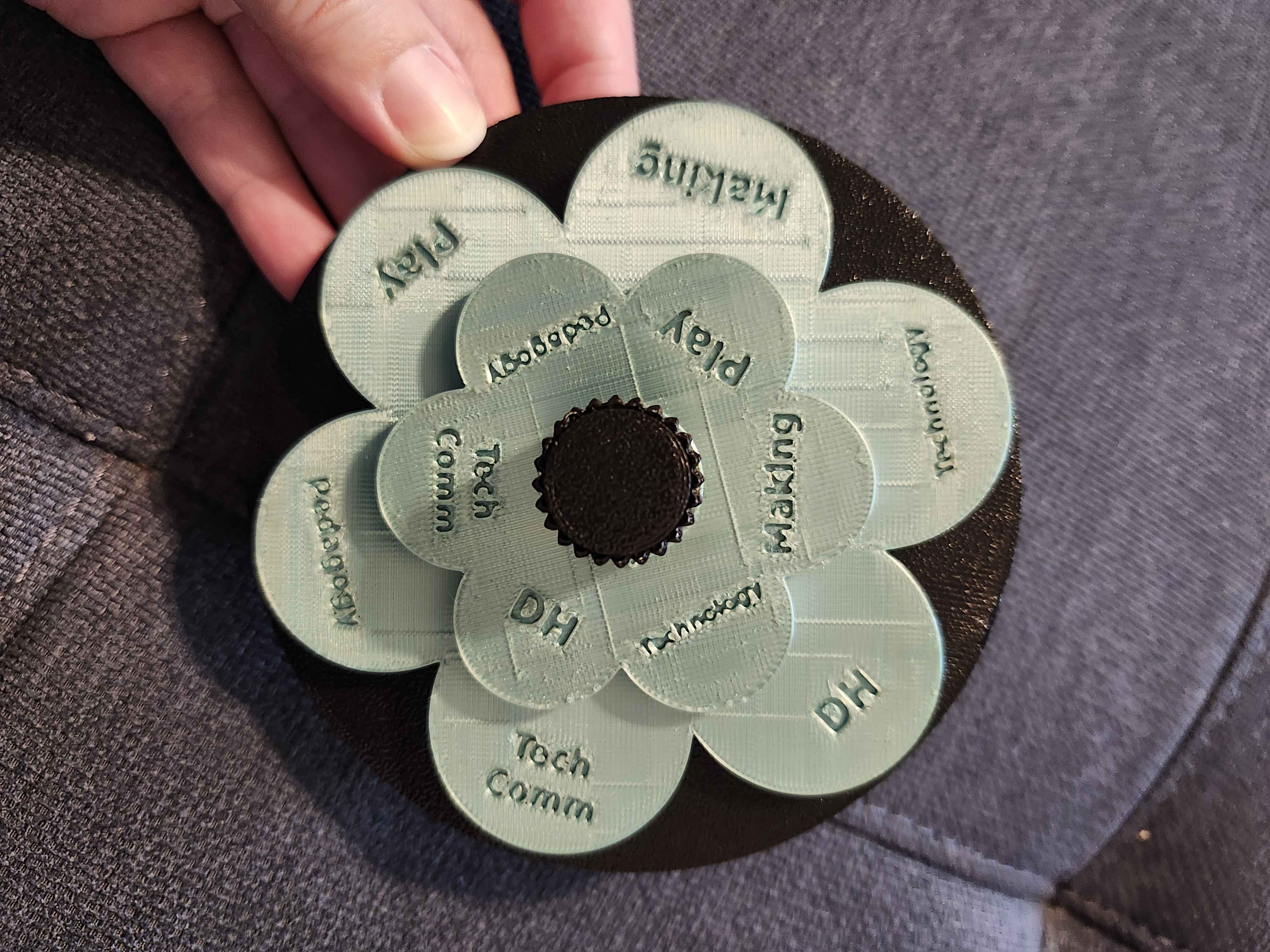 The finished, printed flower spinner held in a hand: two layers of pale green petals with topic words on a black base, with a black knob in the center